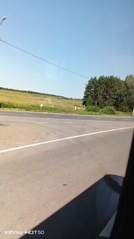 The video shows a view from inside a vehicle, looking out a window at a road with green fields and trees. The sky is blue and clear. A white car passes by in the opposite lane. The video is dated July 2023.