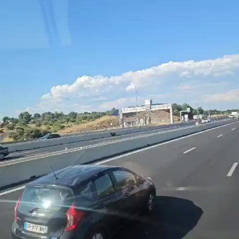 The video shows a view from inside a vehicle traveling on a highway. The road is asphalt with white lane markings. There is a concrete barrier separating the lanes and a hilly, dry landscape with scattered vegetation on the right side. The sky is blue with scattered white clouds. Several cars are visible on the highway, some in front of the vehicle and some in other lanes. The vehicle appears to be moving at highway speeds.