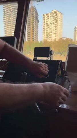 The video shows a view from inside a boat, looking out through a window. In the foreground, a person's arm and hand are visible, resting on a wooden surface. The person appears to be holding a steering wheel. Behind the person, there is a console with various instruments and screens. Outside the window, several tall buildings are visible, along with trees and a body of water. The sky is clear and blue, suggesting it is daytime. The overall impression is of a person navigating a boat in an urban setting.