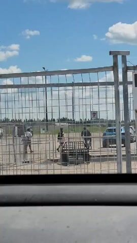 A wire fence runs horizontally across the foreground, partially obscuring the scene behind it. A group of individuals are visible through the fence. On the left, a person in a white shirt and dark pants walks away from the camera. In the center, a person in a striped shirt stands near a dark-colored cart or cage. To their right, a person on a bicycle is moving. Further right, another individual in dark clothing stands. In the background, there are vehicles and buildings under a bright, partly cloudy sky.