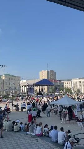 A wide shot shows an outdoor public square with a large crowd of people gathered under a clear blue sky. In the background, there are several multi-story buildings, one of which has a large blue awning over a stage set up in the square. Several tents are also set up in the square, and some people are sitting on steps in the foreground. The scene appears to be a festival or outdoor event.