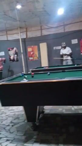 The video captures a scene in a casual recreational setting, likely a pool hall or bar, where three men are playing pool on two tables. The room is lit by overhead lights. Two posters featuring beer advertisements adorn the back wall. 

The pool tables are black with green felt. One man, wearing a yellow shirt, is lining up a shot on the nearest table. Another man in a white T-shirt and a baseball cap observes from the other table. A third man, dressed in a light-colored patterned shirt, walks behind the pool table. 

The flooring is partially visible and appears to be tiled. The background suggests a relaxed, unpretentious environment. The sound in the video appears to be dominated by music.