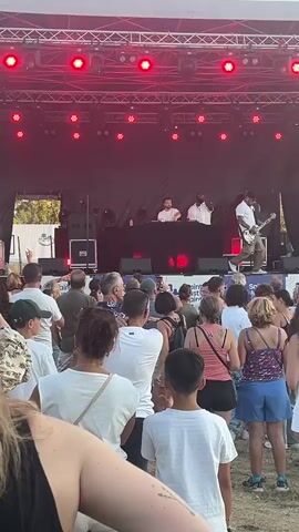 A live music performance is taking place outdoors on a stage. The stage is illuminated by red spotlights. On the stage, there are three men visible. Two men are standing behind a DJ booth, and one man is sitting on a stool playing a guitar. A crowd of people is gathered in front of the stage, facing the performers. The sky is clear and blue. The overall atmosphere appears to be a festival or outdoor concert.