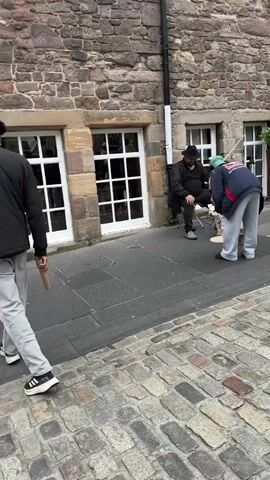 In a cobblestone street in Edinburgh, Scotland, a street performer, a man in a black hat and leather jacket, is seated playing a cello. A small white dog stands beside him, near a basket with items inside. Another person, dressed in a black hoodie with white lettering that reads "SUML" on the back and white cargo pants, stands watching the performance. The building behind them is made of rough-hewn stone with multiple windows. The time of day appears to be daylight, and the weather seems mild. The scene captures a moment of street art and entertainment in a historic urban setting.