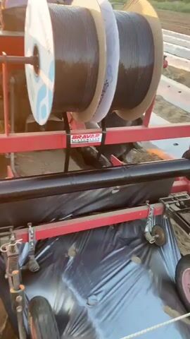 The video shows a close-up view of agricultural equipment being used in a field. Two large spools of black plastic material are mounted on a red frame. A black plastic sheet is being unrolled and laid down on the soil. The equipment has wheels and appears to be attached to a tractor, which is partially visible in the background. The scene is outdoors, with rows of plants or tilled soil visible in the distance. The lighting suggests it is either early morning or late afternoon. The overall activity appears to be the laying of plastic mulch for agricultural purposes.
