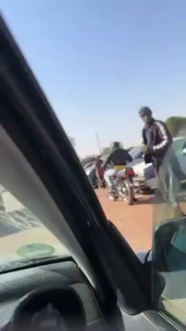 The video is shot from inside a car, looking out through the driver's side window. The car appears to be stuck in traffic. Outside the car, several people are visible along with other vehicles. Initially, a person in a red and black striped shirt stands near the car. Later, a man in a blue shirt and light-colored shorts appears and stands near a blue car, seemingly engaged in some activity around the vehicle. The sky is clear and blue. Power lines stretch across the sky above the road. The environment suggests a busy, possibly congested street in a warm climate.