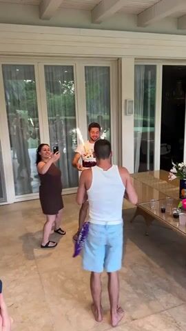 A man in a white tank top and blue shorts dances and smiles while holding his hands up as another man in a white t-shirt holds a birthday cake with sparklers. A woman in a brown dress holds a phone and records the scene. The video is shot outdoors on a tiled patio with glass doors in the background.