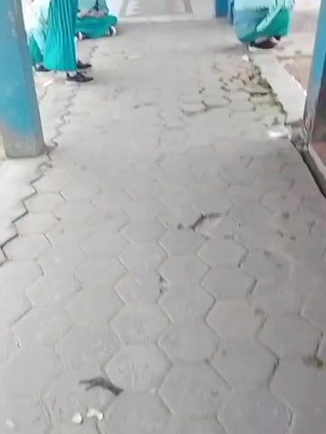 The video shows a hallway with a tiled floor. There are four individuals visible, all wearing light blue hijabs and blue and white checkered uniforms. Two individuals are seated on the ground, one in the foreground and one further back in the hallway. A third individual is standing on the left, facing away from the camera. The fourth individual is squatting on the right side of the frame, with their back to the camera. The hallway has blue columns and a wooden door at the end. Some of the tiles on the floor are cracked and broken.