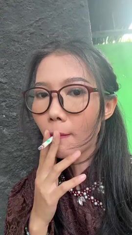 A young woman with long dark hair, glasses, a brown lace top, and a white beaded necklace with silver accents stands facing the camera. She has pink lipstick and makeup on. She is positioned in front of a dark gray wall, with a bright green wall visible in the background to her right. At 0:02, she exhales a plume of white smoke from her mouth, which dissipates by 0:04. She remains stationary, looking at the camera throughout the video.