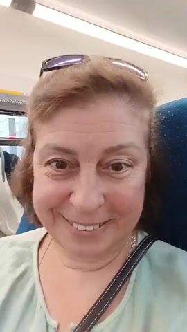 A first-person perspective video shows a woman with brown hair smiling and looking directly at the camera. She is wearing a light green shirt and sunglasses on her head. The background appears to be the interior of a train carriage, with rows of empty blue and white seats visible. The lighting is bright, and the camera is slightly shaky, suggesting movement. The video pans slightly to the right, revealing more of the train's seating area and a woman with blonde hair sitting in a seat further down the carriage, looking at her phone. The woman in the foreground briefly turns her head to the side before returning to face the camera.