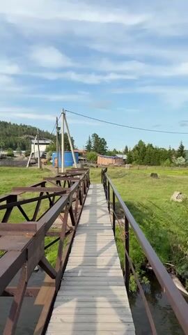 A wooden bridge with a rusted metal frame extends over a small stream. A black bird is perched on the railing of the bridge. In the background, there are several buildings, trees, and a grassy field. The sky is blue with some white clouds. The video appears to be taken on a sunny day.