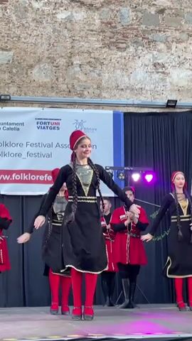 A group of dancers, predominantly female, are performing on a raised stage in front of a backdrop banner. The dancers are wearing traditional folk costumes. The women in the foreground are dressed in black dresses with gold trim and red tights, and red headscarves. They have long braided dark hair. Some dancers in the background are wearing red tunics with black trim and black boots. They appear to be clapping. The banner behind them has logos and text in Catalan and English, including "Ajuntament de Calella", "FORTUNI VIATGES", and "FOLKLORE FESTIVAL ASSOCIATION", and a website address "www.folklorefestival.com". The background wall appears to be made of stone. Stage lighting is visible, with purple and red lights illuminating the scene. The overall setting suggests an outdoor festival or performance.