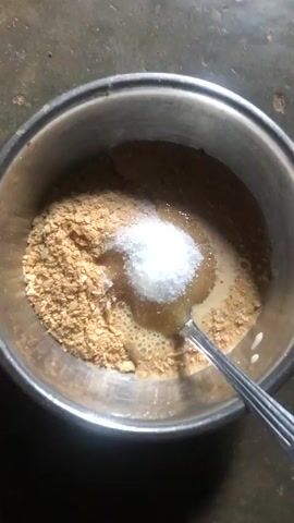 Here's a concise description of the video:

The video shows a metal bowl containing a mix of what appears to be crushed crackers or flakes, a clear liquid (possibly honey or syrup), and granulated sugar. A metal spoon is used to stir the ingredients together, blending them into a slightly gooey mixture. The bowl is placed on a gray surface.
