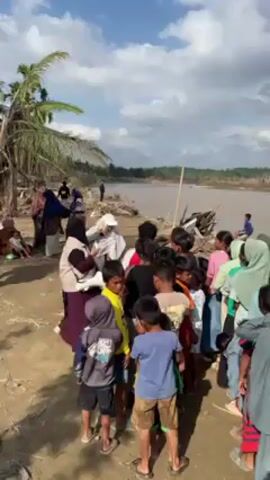 A large group of children, primarily of South Asian descent, are gathered on a muddy riverbank.  They are mixed in age and gender.  Many of the girls wear hijabs.  Adults, also wearing hijabs, stand among them. The children appear to be receiving something from the adults. The environment is a riverbank, with a wide, muddy river in the background and a partially cloudy sky above. Debris is scattered along the shoreline.