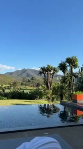 The video shows a view of a swimming pool with mountains in the background on a clear, sunny day. The pool has a dark, tiled bottom. Beyond the pool is a grassy area with trees and various plants, leading to a mountain range. The sky is a bright blue with a few scattered clouds. The camera is positioned close to the pool, suggesting the viewpoint of someone lying on a lounger. There is some indistinct talking in the background, possibly German.