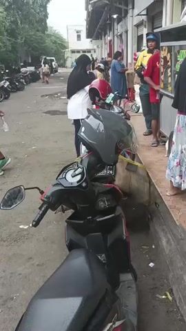 A group of people are gathered outside a food stall. The stall has a sign with Indonesian writing, and appears to be selling snacks. Several motorcycles are parked in the background. The people are standing in line, and some appear to be ordering or receiving food. The scene is outdoors, and the weather appears to be overcast.