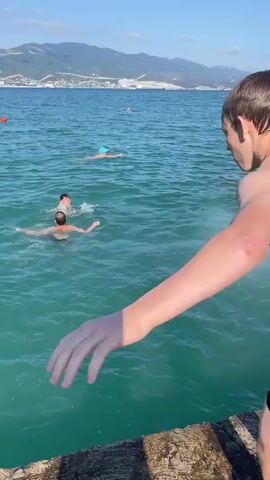 The video shows a bright, sunny day at a coastal location. The primary subject is a young man, shirtless and wearing black swim shorts, standing on a concrete ledge at the edge of the water. He is in mid-leap, having just jumped or about to jump into the sea. His arms are extended outwards, and his body is angled downwards.

The water is a vibrant turquoise color, and several other people are visible in it. One person, further out, appears to be on a small flotation device. Two other individuals are swimming closer to the jumping spot, and a person wearing a blue cap is further out. The activity is clearly swimming and recreational jumping into the sea.

In the background, a coastline is visible with hills or mountains rising behind it. There are structures on the coastline, including what appear to be industrial cranes and buildings, suggesting a populated or industrial area near the water. The sky is a clear blue, indicating pleasant weather. The overall mood appears to be one of fun and summer activity. No text is visible in the video. The timestamp "00:00" to "00:04" is overlaid on the video. The location context provided is Sadovyi, Russia.