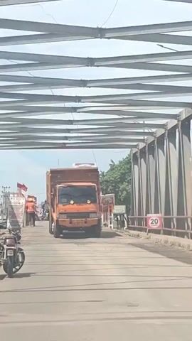 An orange dump truck is driving under a steel truss bridge. The truck has two headlights and a red cargo bed. To the left of the truck, a motorcycle is parked, and two people in orange vests are walking. A red and white flag is visible on the left. To the right of the truck, a speed limit sign indicates 20. The road appears to be paved.