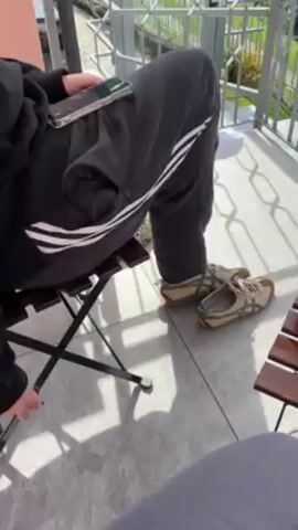 The video shows a balcony view of a lake and mountains. There are two people sitting at a small wooden table. One is wearing gray sweatpants and Adidas shoes, and is holding a phone. The other is wearing gray pants and white sneakers. There are two cups of coffee on the table, as well as an ashtray with cigarette butts. Clothes are hanging on the balcony railing.