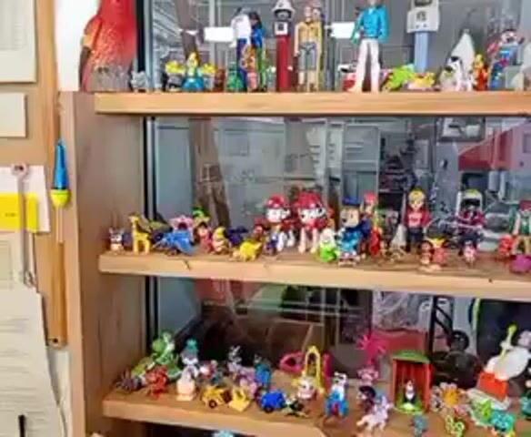 The video captures a detailed view of a three-tiered display cabinet filled with a vast collection of small figurines and collectibles. The camera executes a slow, horizontal pan from left to right, revealing the items on each shelf.

The top shelf features larger figures, including a distinct red bird statue on the far left. Other notable items include several humanoid figures, one resembling a spaceman, an elephant figurine, and various decorative objects, with a blue clock-like piece visible towards the right.

The middle shelf is densely populated with numerous smaller character figurines. A significant portion of this collection appears to be "Paw Patrol" characters, such as Marshall, Chase, Rubble, and Skye, alongside other colorful animal and human-like figures.

The bottom shelf also showcases a diverse array of small, vibrant collectibles. These include figures resembling garden gnomes, mythical creatures, miniature houses or structures, and more cartoon characters. A distinctive figure with pink hair and a white plush rabbit-like creature can be seen among the items.

The setting is an indoor space, likely a shop or a personal collection display, located in Kabile, Bulgaria. Reflections in the cabinet's glass suggest a larger room or commercial area behind the display. The lighting appears consistent, implying an artificially lit environment. No specific activity or event is taking place beyond the filming of the collection, and no discernible text is visible on any of the items or the display unit.