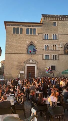 The camera pans across a busy outdoor plaza in Italy. The plaza is surrounded by old stone buildings, and a number of people are sitting at tables in front of an establishment called “Ristorante Bacco.” Most people are sitting and chatting, with drinks and snacks on their tables. Some people are standing, talking, and walking around. The scene is bathed in the warm golden light of a late afternoon sun. Above one of the building’s windows are two red and blue flags. There are a few balloons in the background. Overall, the scene captures a lively and social atmosphere on a pleasant day.