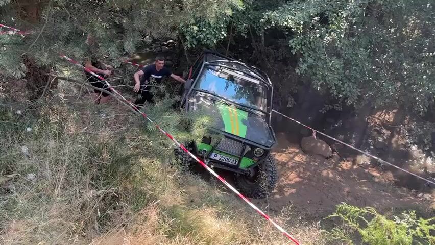 This video shows a black off-road vehicle, possibly a modified Suzuki Samurai, with green and yellow stripes on the hood and a roll cage, attempting to navigate a muddy, wooded terrain. Two men are visible in the frame. One man, wearing a black t-shirt and dark shorts, is on the left side of the vehicle, appearing to be pulling on a rope or strap attached to the vehicle, possibly helping to pull it out of a difficult spot. The second man, wearing a dark t-shirt with white lettering and dark shorts, is standing closer to the vehicle's rear, also seemingly involved in the recovery effort. The vehicle is angled on a muddy slope, with its tires digging into the dirt. Red and white caution tape is strung across the scene, indicating a course or boundary. The environment is overgrown with trees and vegetation, and a small stream or water channel is visible in the background. The overall impression is of an off-road event or challenge where participants are navigating difficult terrain.