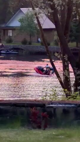 The video shows three people riding a red jet ski in a lake. The water is shimmering in the sunset. A group of people stand on a dock in the background near a couple of parked jet skis. The background also features some cabins and lots of trees.