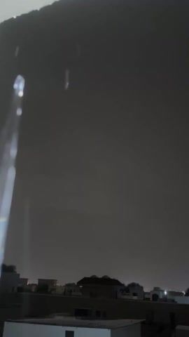 The video shows an exterior shot of a rainy night scene from under a shelter or overhang.  The sky is dark gray and rain is falling.  In the distance, a row of low buildings is visible, with faint lights from within.  The camera is mostly stationary.