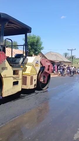 A steamroller is compacting asphalt on a road, with a crowd of people gathered on the side of the road watching. Some people are on motorcycles, and there are buildings and vegetation in the background. The sky is blue with some clouds.