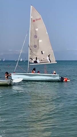 A small sailboat with three passengers, two children and one adult, glides on the water under a clear blue sky. The sailboat is light blue with a tan sail. A person in an orange life jacket is dangling their legs off the back of the boat. In the background, other sailboats can be seen on the horizon. The water is calm and clear.