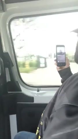 A person is sitting inside a white van and recording a video of the scenery outside the window with a smartphone. The person is wearing a black jacket with a yellow design and blue jeans. The background shows a blurry view of trees and buildings passing by. The phone is held up to the window. There is loud music playing.