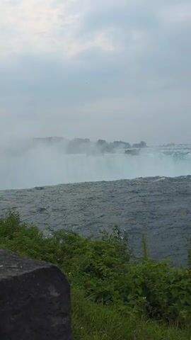 The video shows a sweeping view of a large waterfall, likely Niagara Falls, on a cloudy day. In the foreground, lush green foliage and grass line the edge of the scene. The water of the river is a deep, choppy blue. The waterfall itself is a powerful cascade of white water, with a thick mist rising from its base. In the distance, a small island covered in trees is visible in the middle of the river. Further in the background, a line of trees forms the distant shore. Towards the end of the video, people can be seen standing on a walkway, looking out at the falls and taking pictures. The sky is overcast and gray.