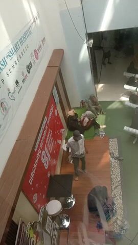 This video provides an overhead view of a public indoor space with several people present. The scene appears to be a common area, possibly a shopping mall or a large building lobby. The flooring is a mix of light-colored tiles and wooden planks.

In the upper left section of the frame, there is a large glass partition, reflecting some of the activity. Beyond this partition, multiple rows of white chairs are visible, and several individuals are seated or standing.

The central and right portions of the frame show the main area of interest. Several people are gathered on the floor, some sitting and others lying down. One person is lying on their back on a green mat, wearing a light-colored shirt and pants. Another person is seated next to them, wearing a headscarf. A third person is lying on their side, also on a mat. There are bags and personal belongings scattered around this group.

A prominent red banner is laid out on the floor with text in what appears to be Indonesian, indicating a celebration or event, possibly related to the "REPUBLIK INDONESIA". The banner features the number "81" in large font.

Other individuals are visible in the background, some standing and others sitting. Near the red banner, there are some reflective silver objects, possibly bowls or containers, and white circular objects. Further to the right, more people are seated on the floor, and footwear is visible. The lighting suggests an indoor environment with overhead illumination. The overall impression is a casual gathering or rest stop within a public building.