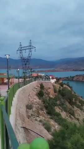 The video shows a view of a lake with surrounding land, likely a reservoir, under an overcast sky. In the foreground, there's a green railing and some vegetation. A tall electrical transmission tower is visible near a building complex with red roofs. The camera slowly pans to the right, showing more of the lake and the hilly, arid landscape around it. The water has a teal color. In the distance, mountains are visible beneath the cloudy sky. As the camera continues to pan, a road and a tunnel can be seen along the shoreline.