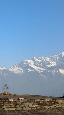 The video showcases a magnificent view of snow-capped mountains, identified as part of the Annapurna range, under a clear blue sky. The initial frame features a low stone wall in the foreground, with the vast, hazy mountain range dominating the background. As the video progresses, a woman with long blonde hair, dressed in dark attire, appears on a grassy hill, gazing at the majestic peaks. She then walks across the scene. Subsequently, another person, also in dark clothing and potentially carrying a backpack, is seen standing on a different part of the hill, facing the mountains, seemingly lost in admiration. A simple wooden post is visible in the midground in one shot. The setting is a serene mountain landscape, likely a viewpoint in Lwang Ghalel, Nepal, offering breathtaking vistas of the Himalayas on a bright, clear day. The people appear small against the grandeur of the mountains, emphasizing the scale of the natural scenery.