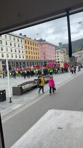 The video shows a group of people gathered in a city square. Many are wearing yellow vests and holding signs with red backgrounds. There is a red tent or canopy in the center of the group. Some people are standing around, while others are walking. Two women, one in a yellow jacket and one in a pink jacket, are walking away from the group. The square is surrounded by colorful buildings. The video is taken from inside a vehicle, looking out through a window.