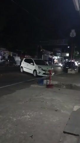The video shows a white car parked on the side of a street at night. There are other vehicles and buildings in the background. The streetlights are on, illuminating the scene. The car appears to be a hatchback. There is a red cone in front of the car. The road is paved, and there is a sidewalk in the foreground. The sky is dark.