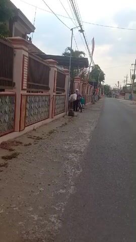 A person on a bicycle is seen from behind and to the side, walking alongside a street. The person is wearing a white hat, a striped shirt, and dark pants. They are carrying a large, white bag on the back of the bicycle. The person is walking on a dirt and gravel path next to an asphalt road. A fence and a building are visible to the left of the path. Indonesian flags are displayed on poles along the fence. The scene appears to be in a residential area.