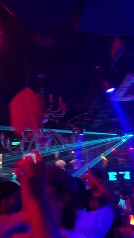 The video shows a crowded nightclub or party scene with many people dancing under vibrant pink, blue, and green strobe lights and laser beams. A large white chandelier with red, string-like decorations hangs from the ceiling. People are moving their bodies and raising their arms, some looking towards the front. The camera is held by someone within the crowd, capturing the energetic atmosphere. Loud music and cheers are audible.