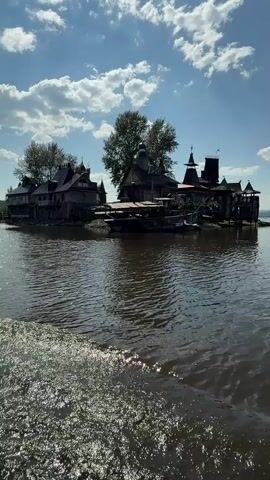 The video captures a view from a boat moving on a body of water. In the foreground, choppy water with white foam indicates the boat's wake. In the midground and background, an island is visible, densely populated with a cluster of wooden structures. These structures appear to be multi-story buildings with pitched roofs and architectural elements resembling traditional or fantasy-inspired designs, including turrets and balconies. Some of the buildings are connected by walkways or platforms. Several boats or floating structures are docked around the island. Trees are interspersed among the buildings. The sky above is blue with scattered white, fluffy clouds, suggesting a bright, possibly breezy day. The water is a dark, murky brown, reflecting the sky and structures. The overall impression is of an unusual, somewhat secluded settlement on an island. There are no people visible in the frame.
