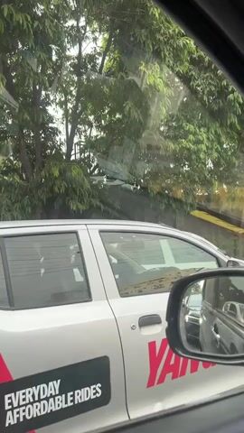 The video shows a view from inside a vehicle, looking out the side window and through the windshield. A white car with a graphic featuring the word "YAMAHA" and the text "EVERYDAY AFFORDABLE RIDES" is visible next to the vehicle. Trees and foliage are present in the background. The view through the windshield shows a traffic jam with several cars, including a white sedan and a dark-colored car. A side mirror of the vehicle the video is being filmed from shows reflections of cars in traffic. The overall impression is of being in a car stuck in traffic on a cloudy day.