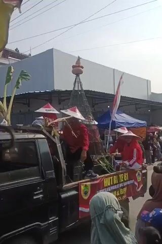 The video captures a parade float featuring several individuals wearing red shirts, black pants, and conical hats. These hats have red trim and appear to be inscribed with white lettering that includes the word "GEMOLONG." The individuals are seated on the bed of a black pickup truck adorned with a large banner. The banner, primarily red and white, displays text related to "HUT RI KE 81," "INDONESIA BERDAULAT ADIL DAN MAKMUR," and "KTNA & BPP KEC. GEMOLONG." There is a large, cage-like structure on the float topped with a round, gourd-like object. Green foliage and what appear to be bananas are also visible on the float. The scene suggests a festive or celebratory event, possibly a local parade in Indonesia. The background shows buildings and tents, indicating an outdoor, possibly urban or semi-urban setting, with other people visible in the periphery. The lighting suggests it is daytime.