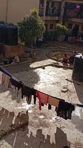 The video shows a sunny courtyard with a cracked concrete surface, surrounded by a multi-story building.  Clothes are hung out to dry on a clothesline that stretches across the foreground. To the left, there are two large black water tanks. Several small trees are scattered throughout the courtyard. In the background, people are gathered in different areas, some sitting and some washing. The video is shot from an elevated position, looking down into the courtyard.