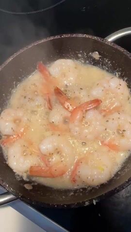A close-up, overhead shot shows shrimp sizzling in a pan with a light-colored, bubbling sauce. The shrimp are pink and white with visible tails. The sauce appears to contain herbs and possibly garlic. The pan has a dark, speckled surface. The background is dark and out of focus, with a partial view of a blue object on the left. Steam rises from the pan, indicating the shrimp are cooking.