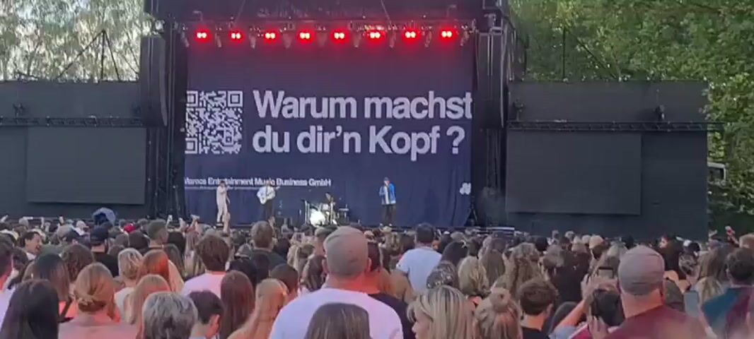 A wide shot captures an outdoor concert stage with a large black backdrop. The backdrop prominently displays a QR code on the left and the German text "Warum machst du dir'n Kopf?" (Why are you worrying?) on the right, in large white lettering. Below the text, smaller text reads "Monca Entertainment Music Business GmbH" and what appears to be a logo. A band of four musicians are performing on stage: a singer in a blue jacket and cap at the front, a guitarist on the left, and a drummer and another musician behind them. Red stage lights are illuminated above the backdrop. On either side of the main stage, two large screens display video content, each also showing a QR code. A crowd of concertgoers is visible in the foreground, looking towards the stage. The environment appears to be a park or open area with trees in the background, suggesting daytime or early evening. The overall scene depicts a live music performance at an outdoor venue.