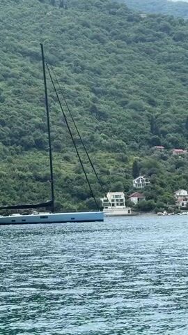 A wide shot shows a light blue sailboat with black sails and masts on choppy blue water. Behind the boat, a densely forested green mountain rises steeply from the shore. Several white houses with red roofs are nestled amongst the trees on the mountainside. The boat is positioned to the left of the frame, and the mountain fills the background.