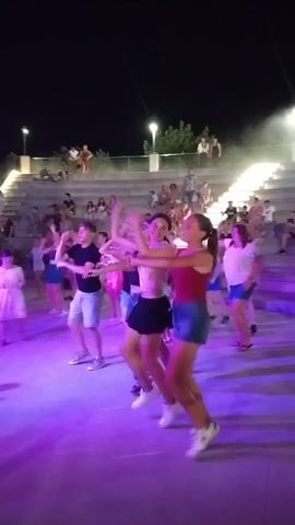 A group of young people is dancing in front of an audience seated in a stadium-like setting at night. The dancers are illuminated by purple and white stage lights. The audience members are visible in the tiered seating area behind the dancers. The environment appears to be outdoors.