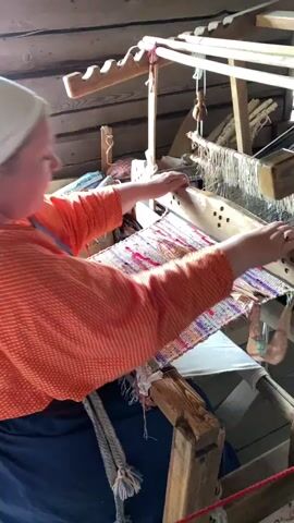 A woman wearing an orange shirt and a white head covering is operating a wooden loom. She is seated and actively engaged in weaving, with her hands positioned over the threads and a colorful fabric emerging from the loom. A pair of scissors lies on the woven material. The background consists of wooden planks, suggesting an indoor, possibly rustic, setting. The loom itself is made of wood and appears to be a traditional weaving device. The woman's facial expression is focused on her task.