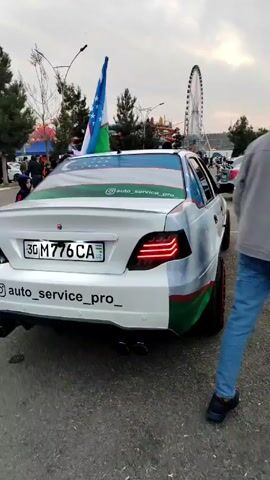 The video shows a white Daewoo Nexia with the colors of the Uzbek flag painted on it and a Uzbek flag sticking out of the rear window.  The car has custom rims with red accents. The license plate number is 30M 776 CA.  The car is parked near an amusement park; a Ferris wheel is visible in the background. Several people are walking around the car. One person touches the car. The Instagram handle "auto_service_pro" is featured on the car.