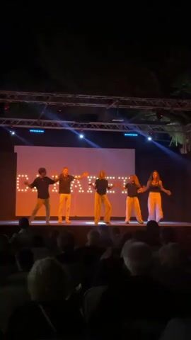 A group of five young people, three males and two females, are performing on an outdoor stage at night. They are all dressed in casual, light-colored clothing, with the males wearing t-shirts and pants, and the females wearing crop tops and wide-leg pants. They are positioned across the stage, facing the audience, and appear to be dancing or performing a choreographed routine, as they are all striking similar poses with their arms extended. Behind them, a large screen displays the word "CAMP" in large, illuminated letters. The stage is lit with blue and white spotlights, casting a bright glow on the performers and the screen. In front of the stage, a dark audience sits in chairs, their faces illuminated by the stage lights. The background shows dark trees and the night sky, indicating an outdoor venue. The overall scene suggests a lively performance in a casual setting.